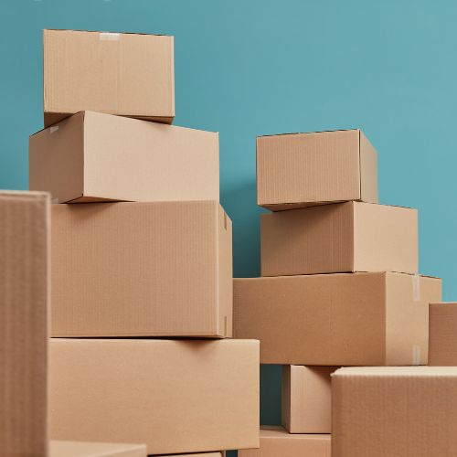 A pile of cardboard boxes against a teal blue background, varying in size and stacked in an irregular fashion, suggesting a theme of moving, storage, or shipping.