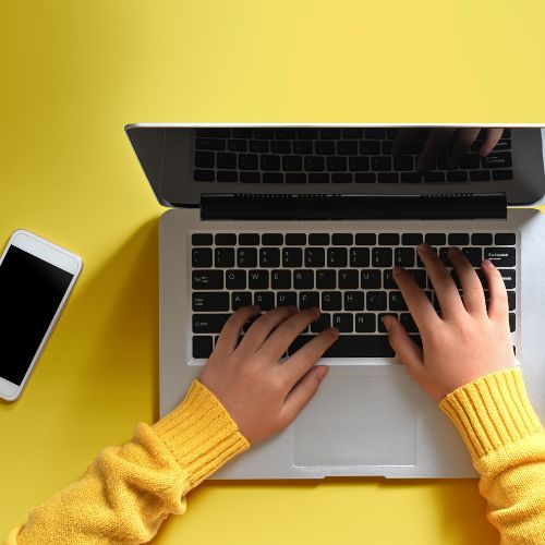 Over-the-shoulder view of a person wearing a yellow sweater typing on a laptop, with a smartphone lying next to it on a yellow surface, representing remote work, online communication, or technology use.