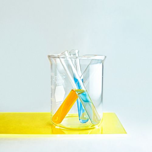 A beaker with a pair of yellow and blue test tubes inside, set against a soft blue backdrop with a yellow base, symbolizing chemical or medical research.